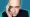 Derren Brown with a serious expression, holding his hand to the side of his face, wearing a dark blazer, against a teal and blue patterned background.